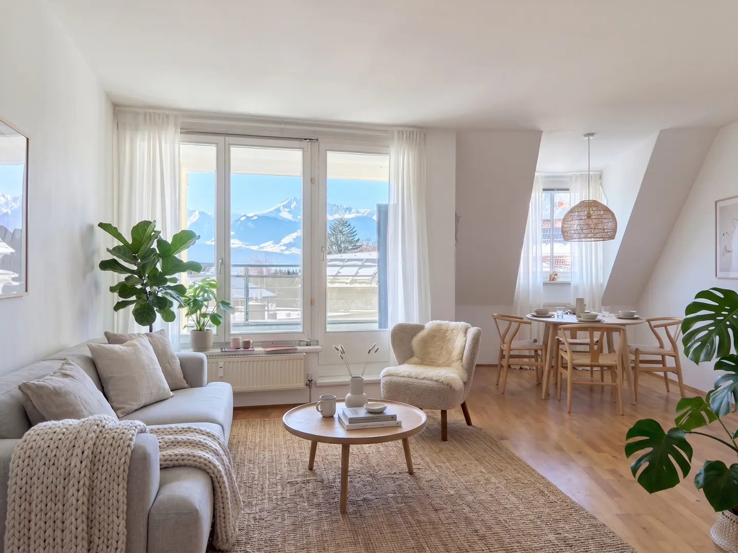 Bright living room with a beige sofa, round table, and dining set. Large windows offer a view of snow-capped mountains.