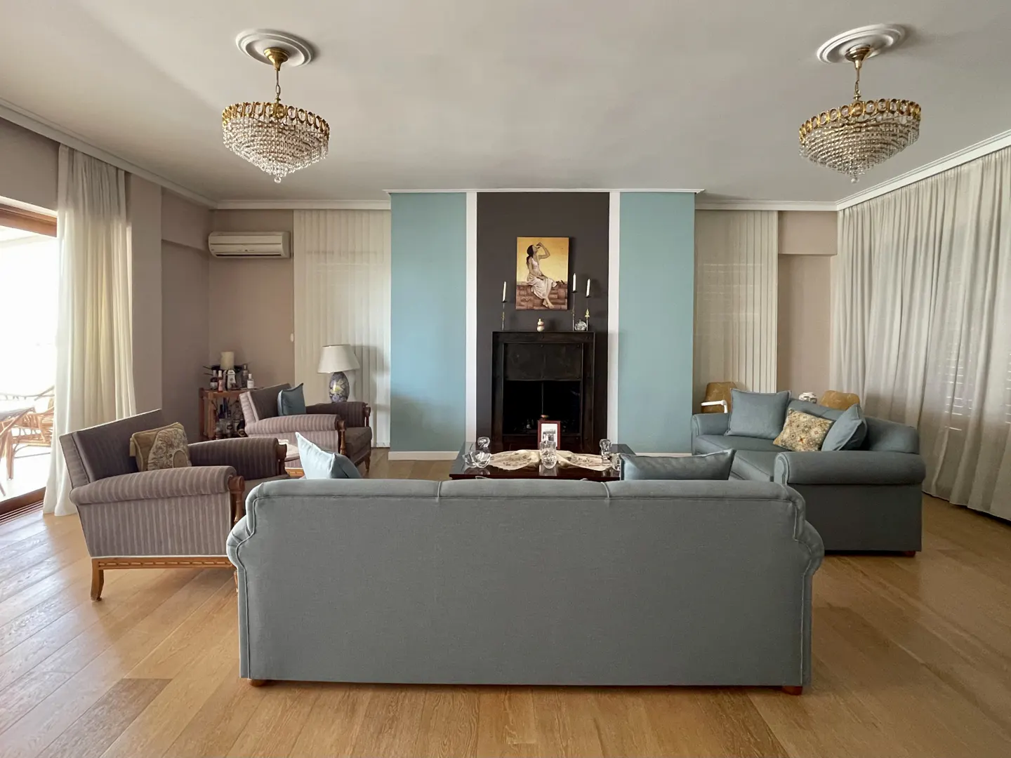 Living room with hardwood floors, gray sofas, armchairs, and a fireplace with a painting above it. Two crystal chandeliers hang from the ceiling.