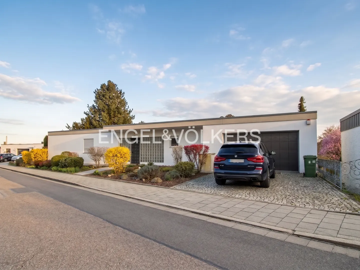 A modern, one-story white house with a dark blue SUV parked in the driveway. The Engel & Völkers logo is visible on the house.