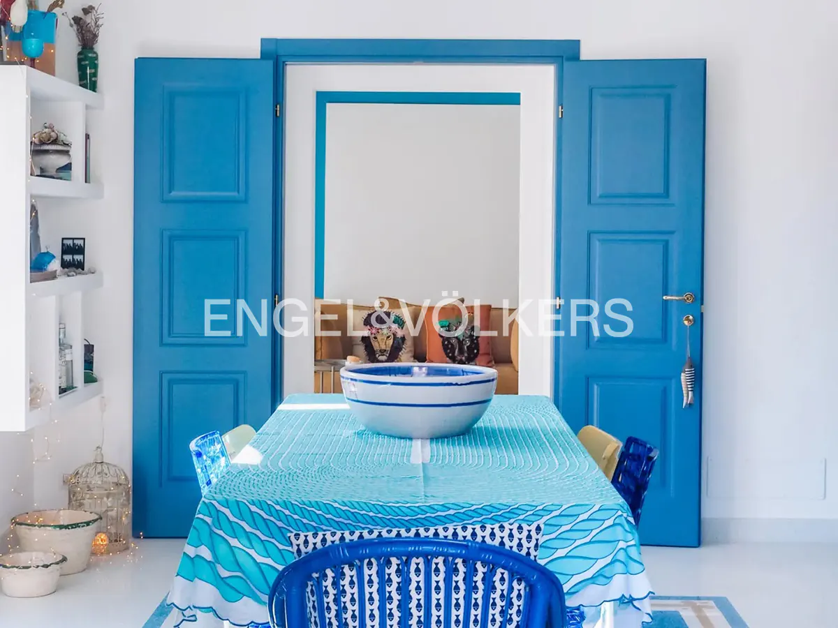 Bright dining room with blue doors, table with blue patterned cloth, and white bowl. Shelves with decor on the left. Engel & Völkers logo.