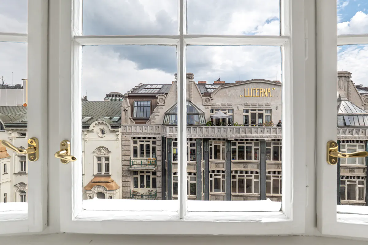 View from a white window with gold handles, overlooking city buildings under a cloudy sky. "LUCERNA" is visible on a building.