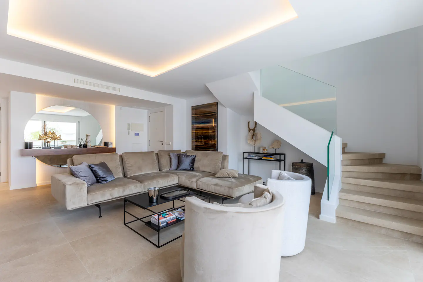 Bright living room with beige sectional sofa, round mirror, white chairs, and staircase with glass railing.