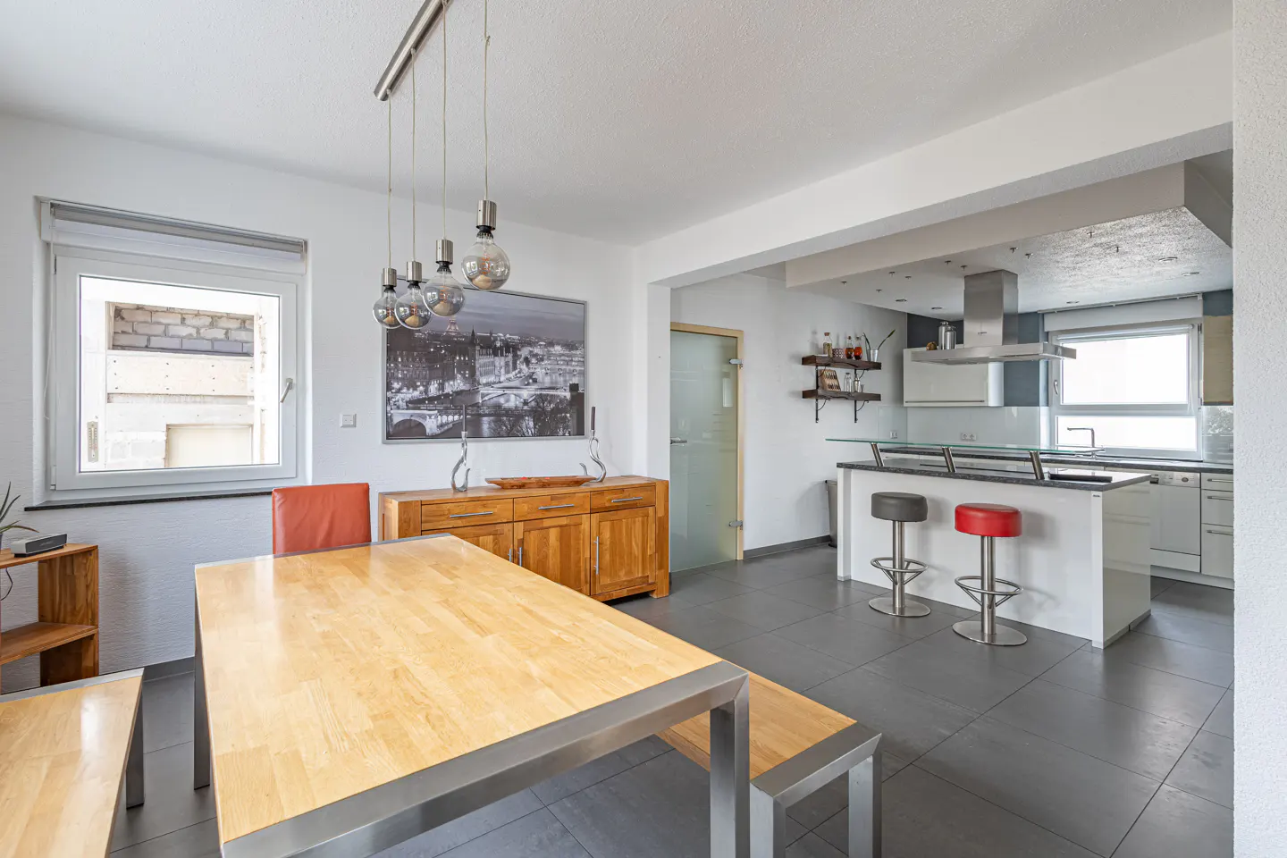 Modern dining area with a light wood table, benches, and a kitchen island with bar stools on a grey tiled floor.
