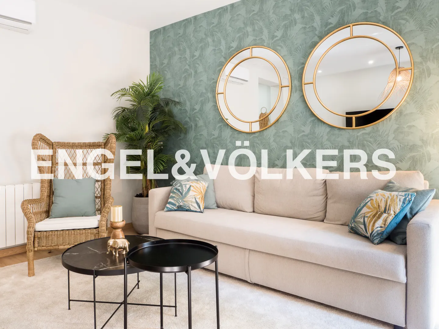 Living room with a beige sofa, wicker chair, black tables, and two round gold mirrors on a green patterned wall.