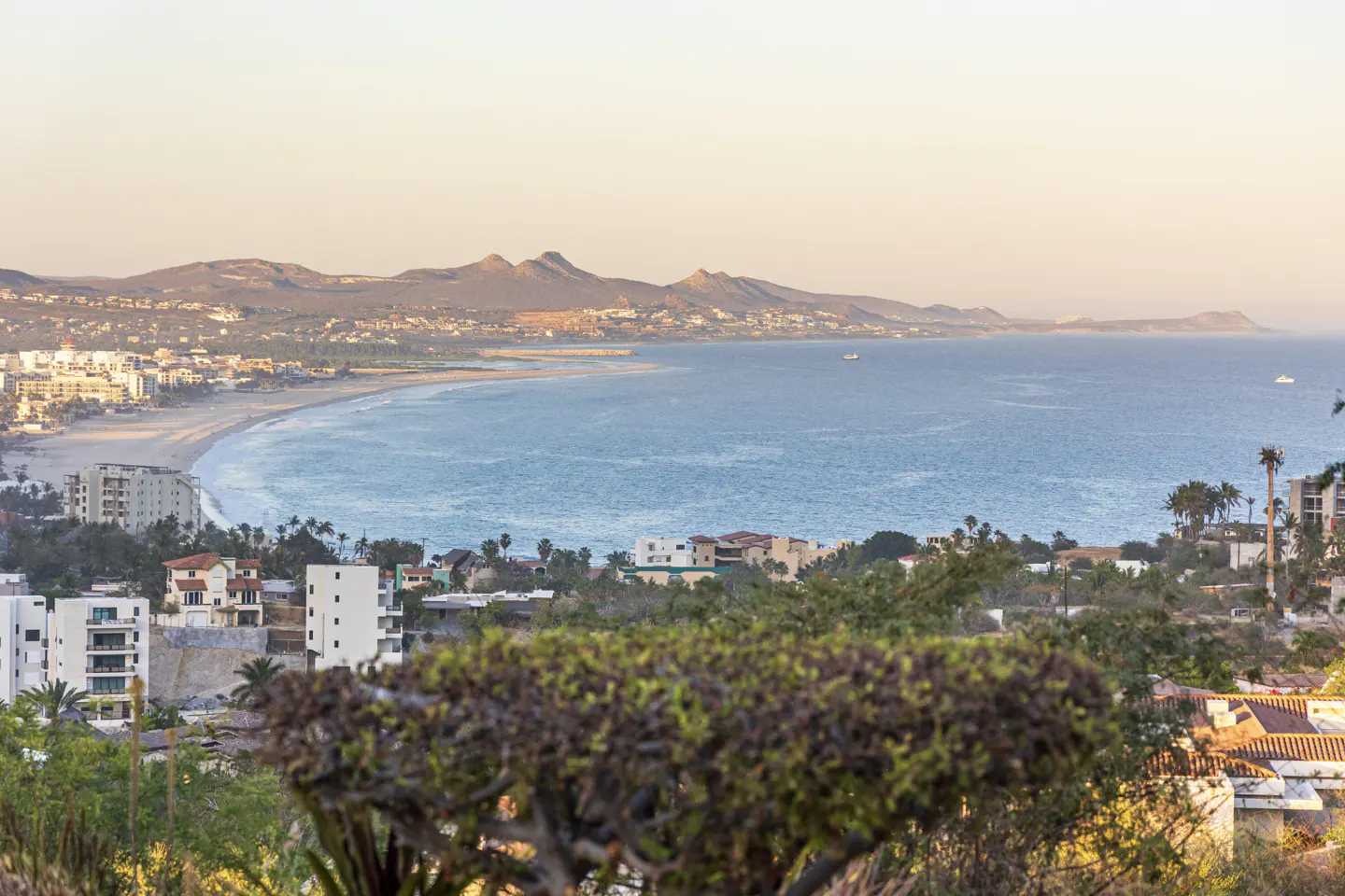 Coastal view of Cabo San Lucas, Mexico, with blue ocean, sandy beach, buildings, and mountains in the background.