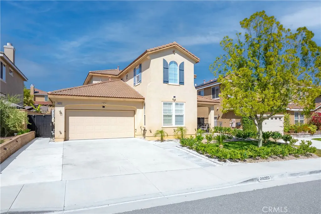 Beige two-story house with a brown roof, blue shutters, and a two-car garage on a sunny day. A green tree is in the front yard.