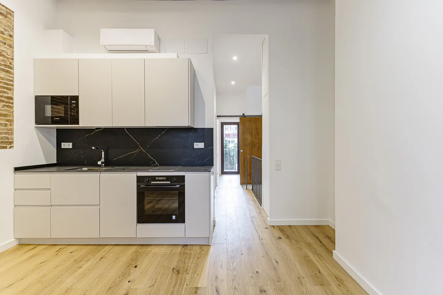 Modern kitchen with beige cabinets, black marble backsplash, and light wood flooring, leading to a hallway with a wood door.