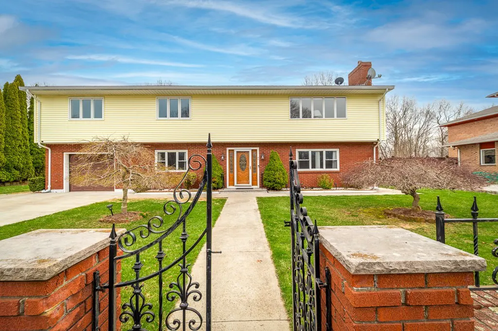 Two-story house with yellow siding and brick bottom, viewed through an ornate black metal gate. A concrete walkway leads to the front door.