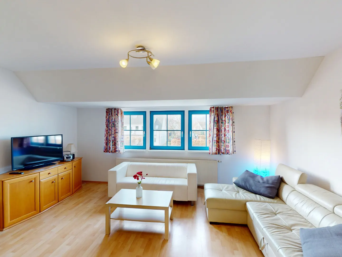 Bright living room with wood floors, white sofas, and blue-framed windows with patterned curtains. A wood cabinet holds a TV.