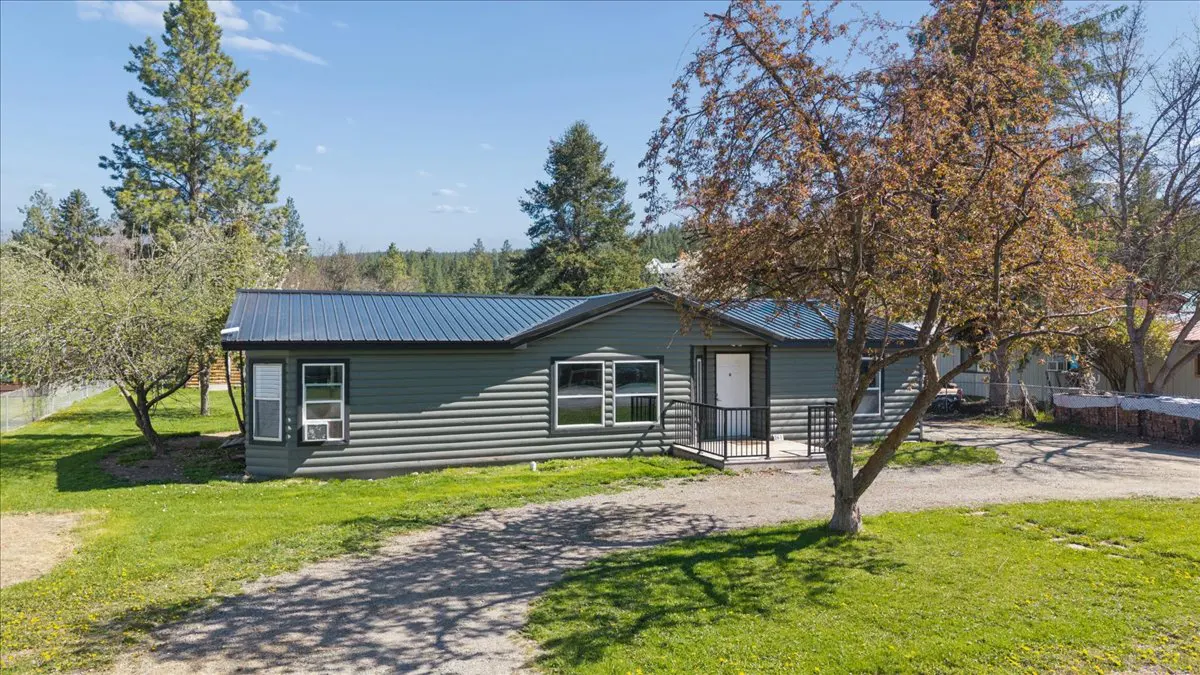 Exterior view of a one-story, green log cabin-style home with a dark metal roof and white trim. A tree is in the front yard.