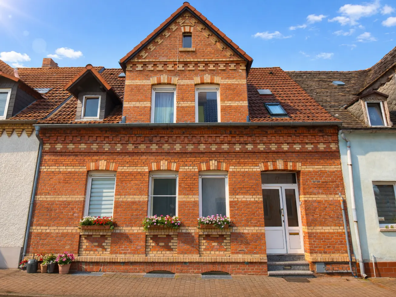 A three-story brick house with white trim, flower boxes, and a red tile roof under a blue sky.