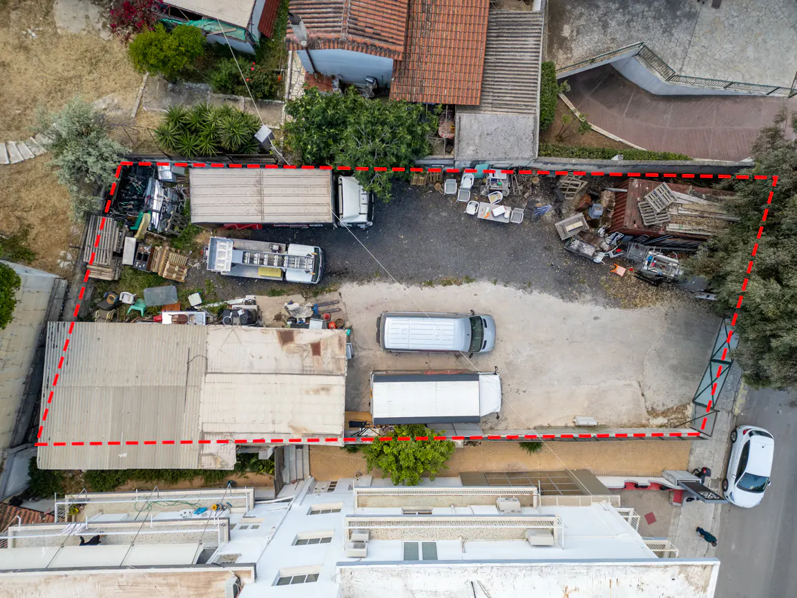 Aerial view of a property outlined in red, featuring trucks, a van, and a building with a gray roof. Debris and greenery are scattered throughout the lot.