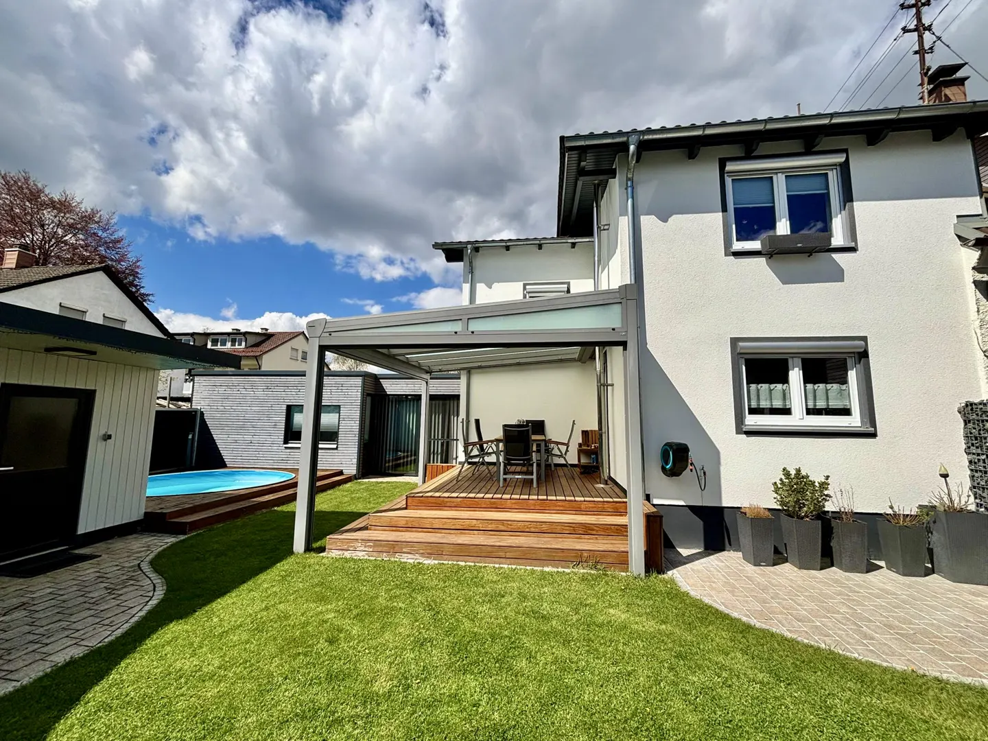 A backyard with a pool, a wooden deck with a table and chairs, and a white house with gray trim.