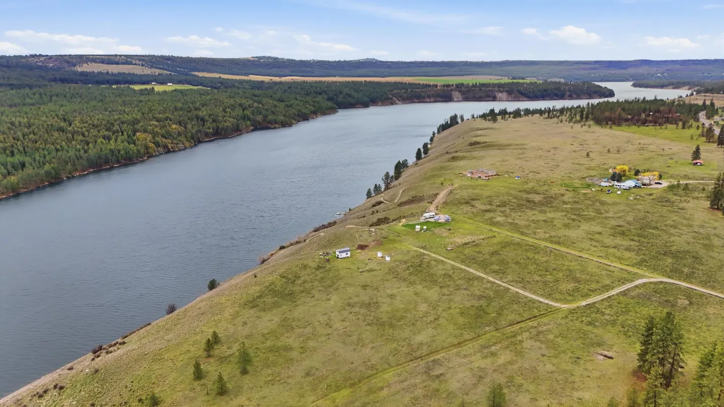 Aerial view of a wide, gray river bordered by green forest and grassy hills with scattered houses under a blue sky.