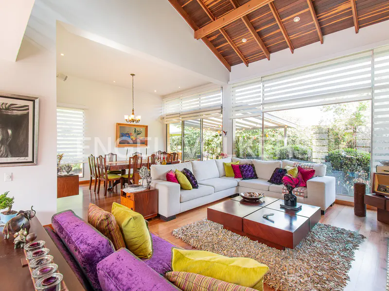Bright, open living room with white sofas, colorful pillows, and a wooden ceiling. Large windows overlook a lush garden.