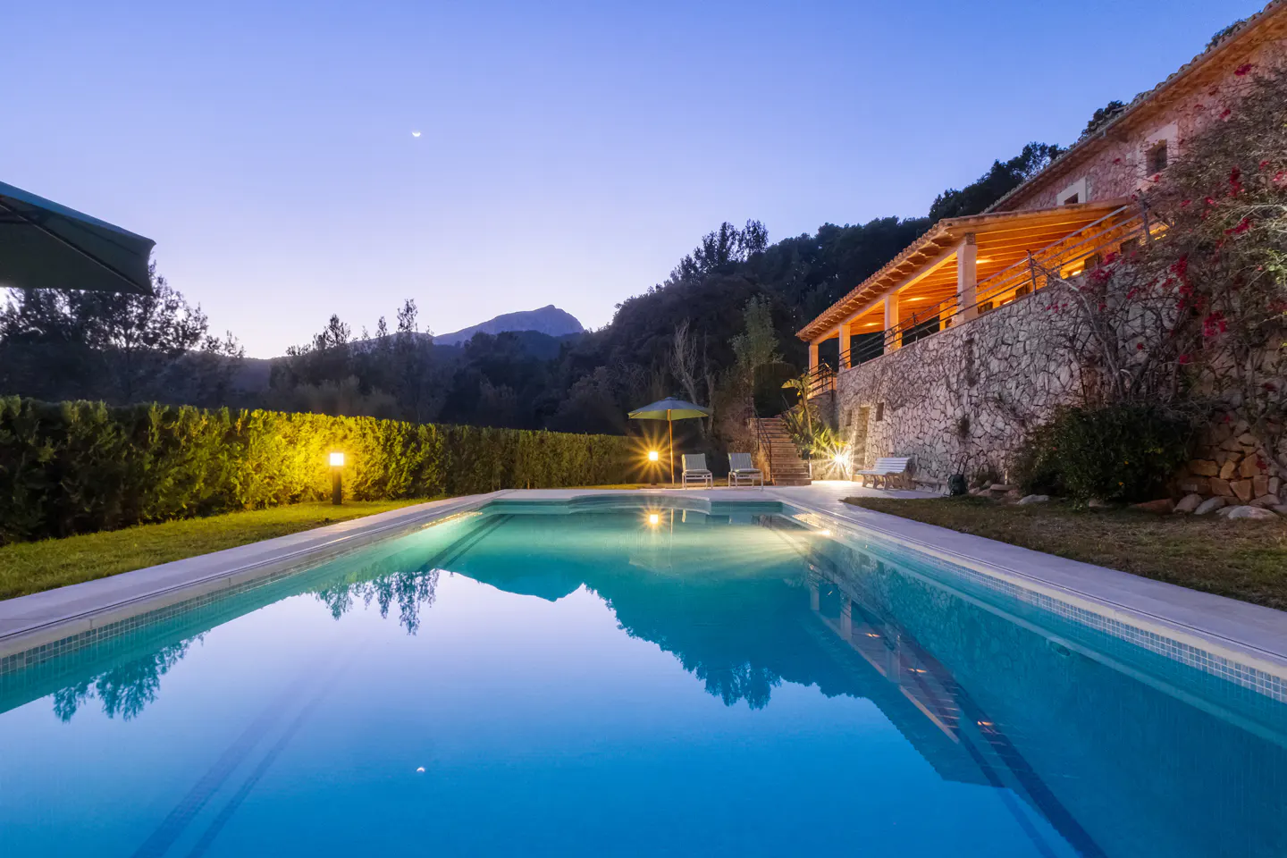 Twilight view of a blue swimming pool reflecting a stone house with a wood-covered porch, trees, and a mountain under a crescent moon.