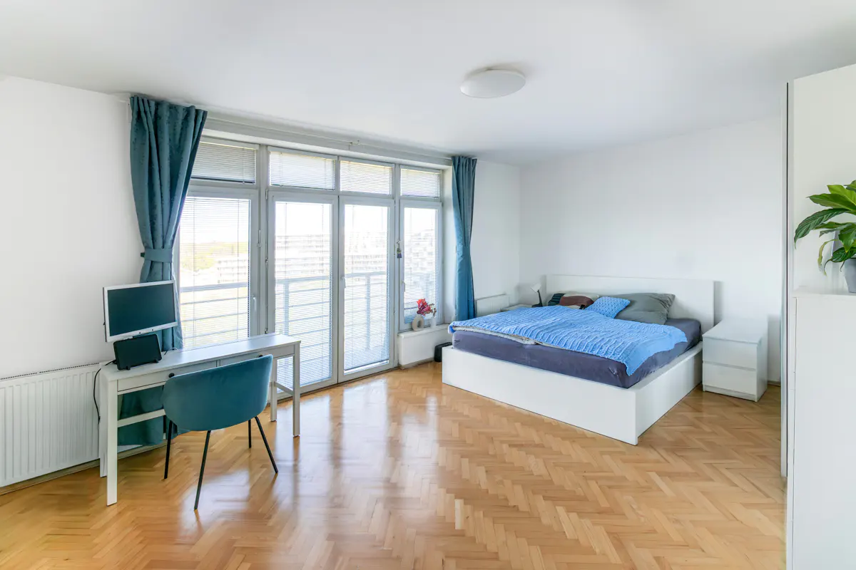 Bright bedroom with white walls, wood floors, and a large window. A desk with a computer and a bed with blue bedding are visible.