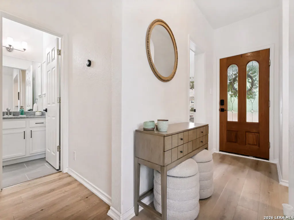 Entryway with wood floors, white walls, and a brown front door with glass panels. A console table with stools sits below an oval mirror. Bathroom visible through an open door.