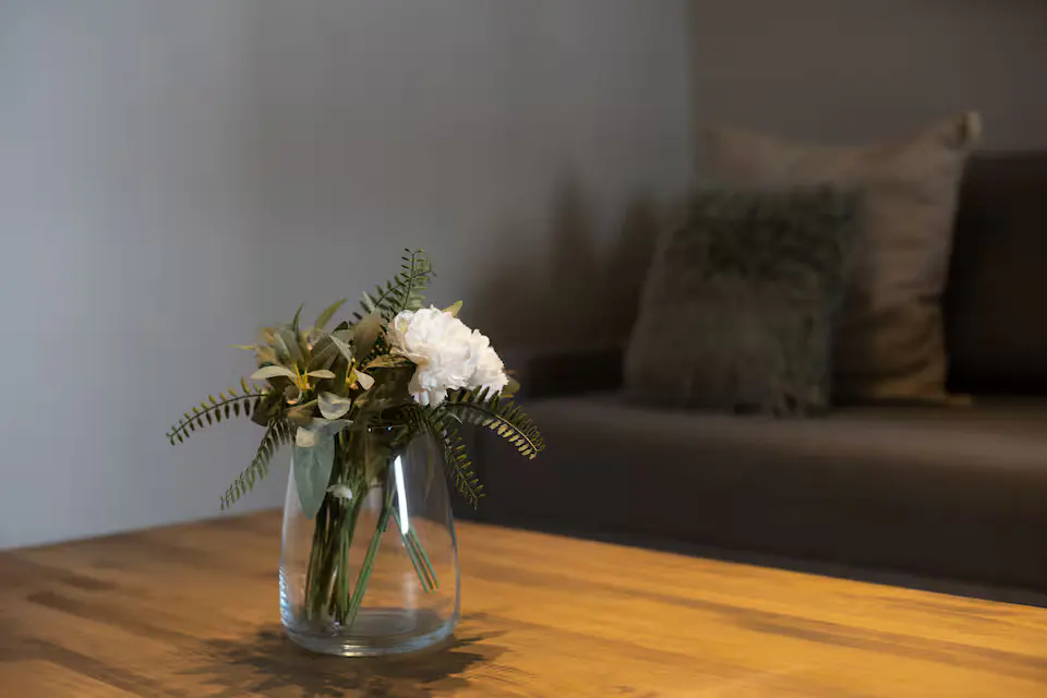 A clear vase with white flowers and greenery sits on a wooden table, with a gray couch and pillows in the background.
