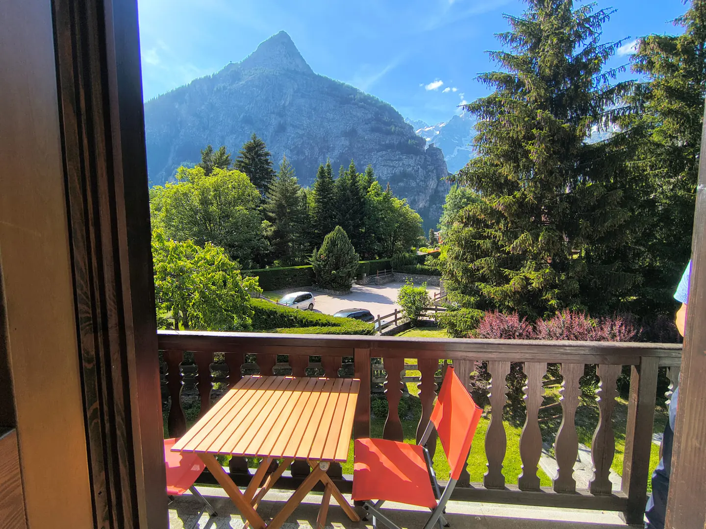 Balcony view with a wooden table and orange chairs overlooking a lush green forest and a towering mountain peak.