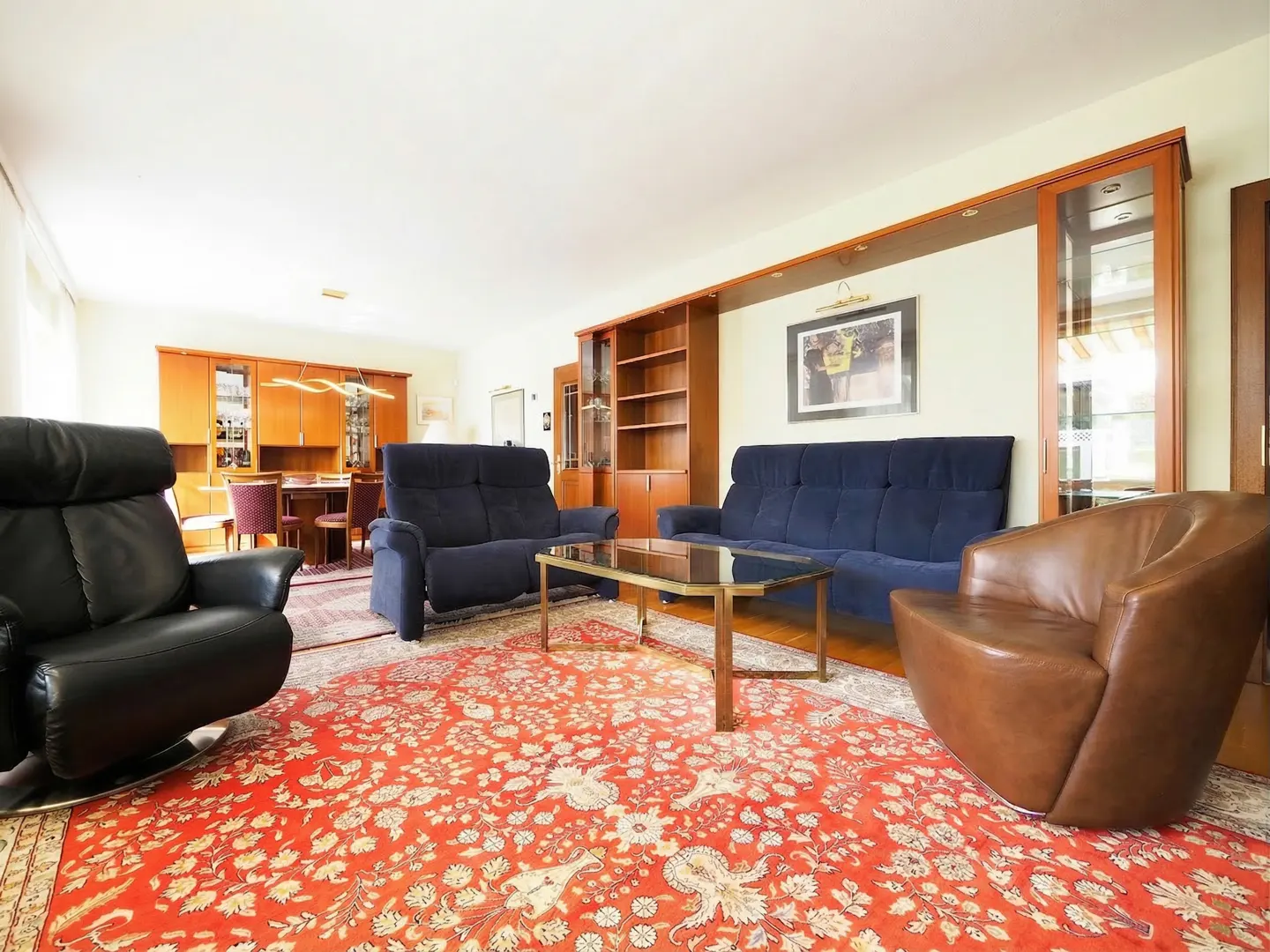 Living room with blue sofas, black recliner, and brown chair on a red floral rug. A glass coffee table sits in the center.