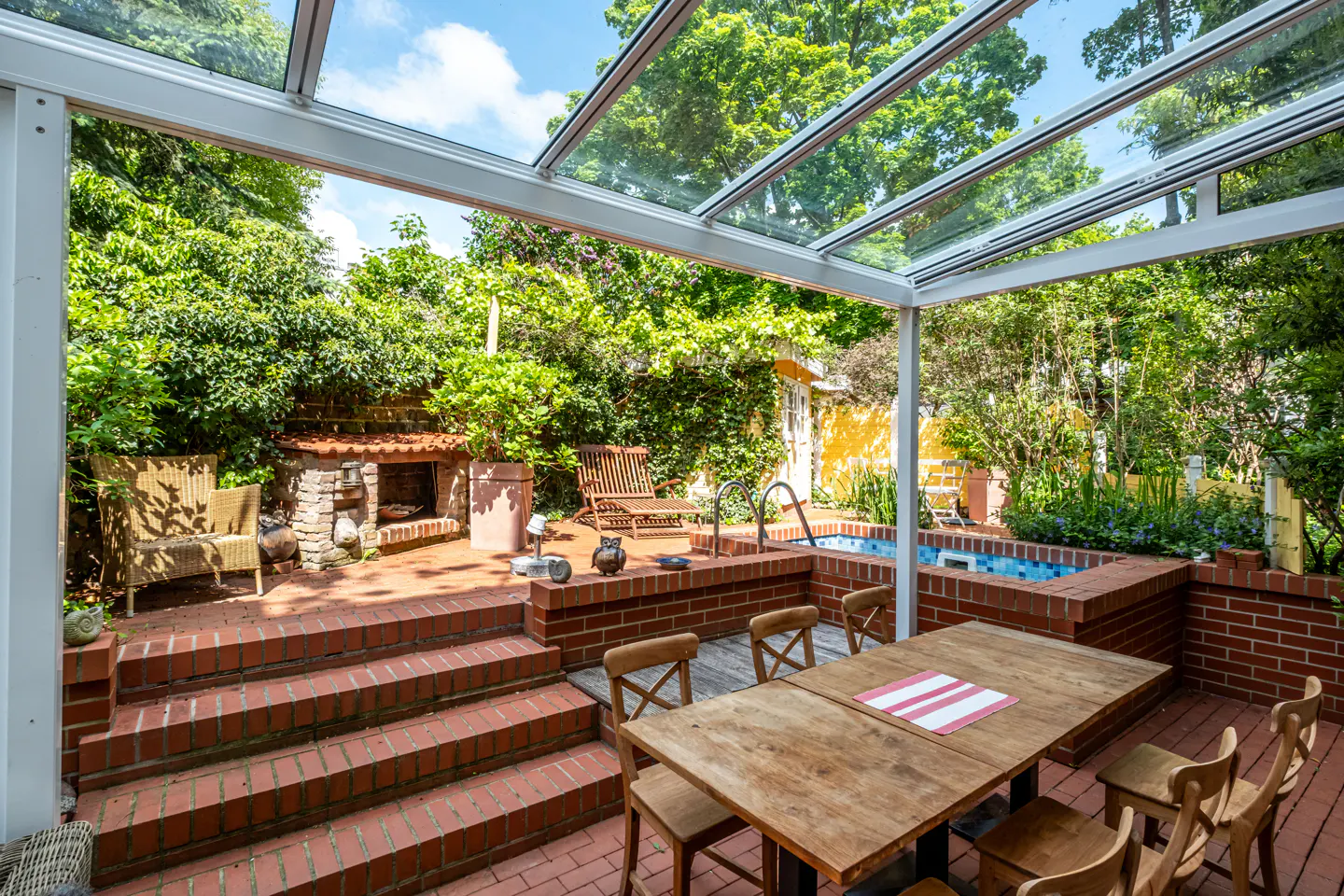 Outdoor patio with glass roof, brick floor, wooden table and chairs, brick fireplace, and small pool surrounded by lush greenery.