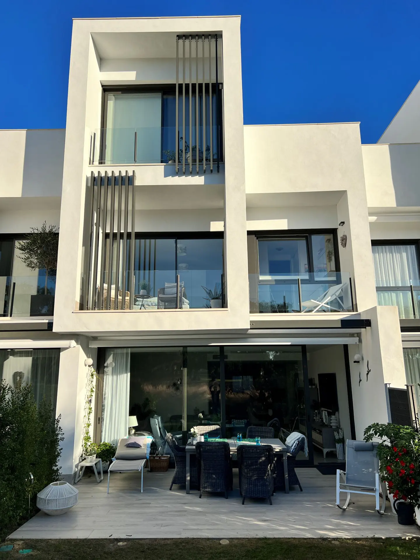 Modern white house with a patio. Wicker chairs surround a table. Lounge chairs are on the patio and balconies. Blue sky.