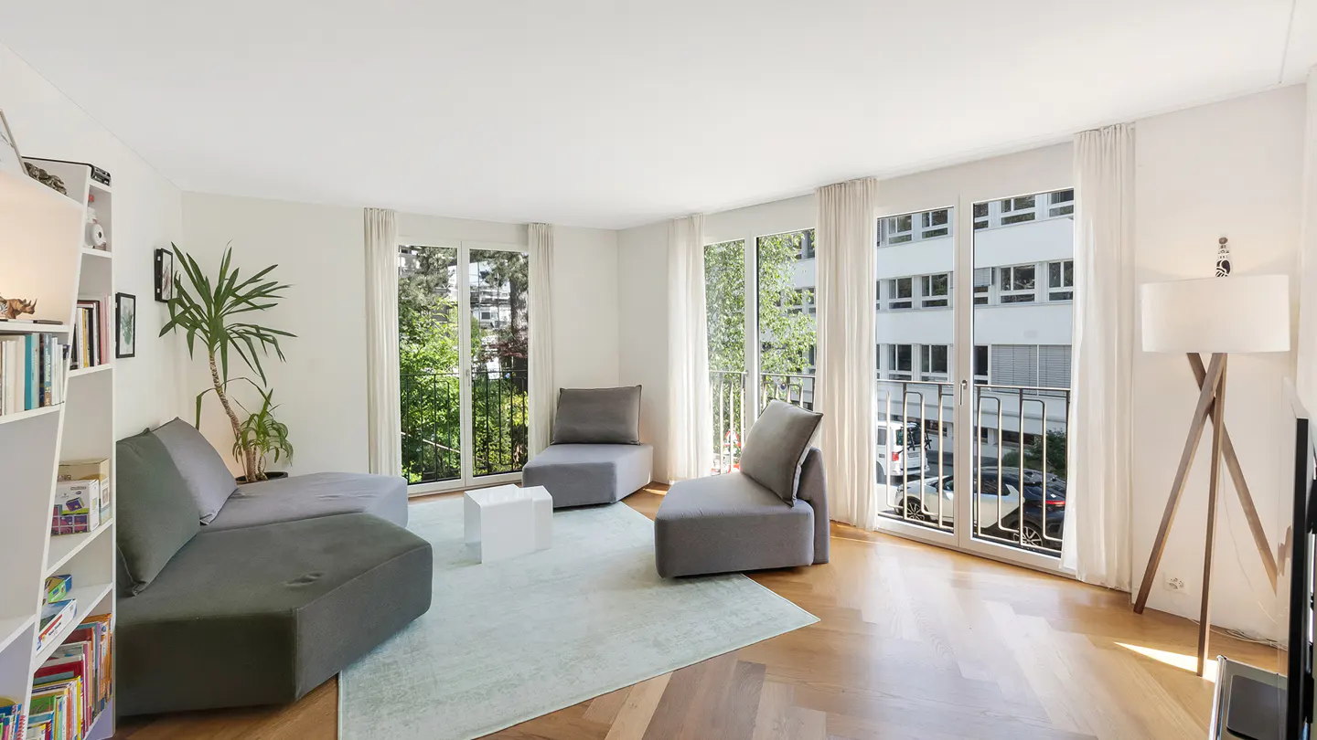 Bright living room with gray sofa, chairs, and light blue rug on wood floor. Large windows with white curtains overlook trees and a building. A bookcase and floor lamp are also visible.