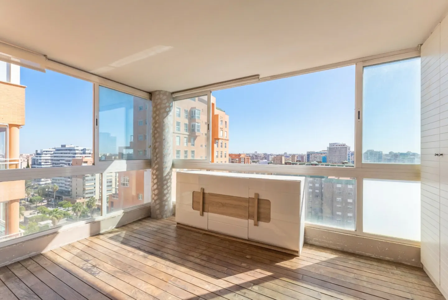 Bright balcony with wood floors and a white cabinet. Large windows offer a city view under a clear blue sky.