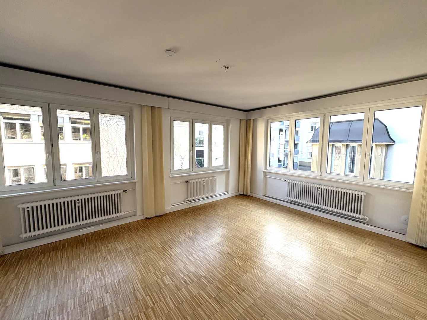 Bright, empty room with parquet floors, white walls, and large windows with white frames and radiators below. Cream-colored curtains frame the windows.