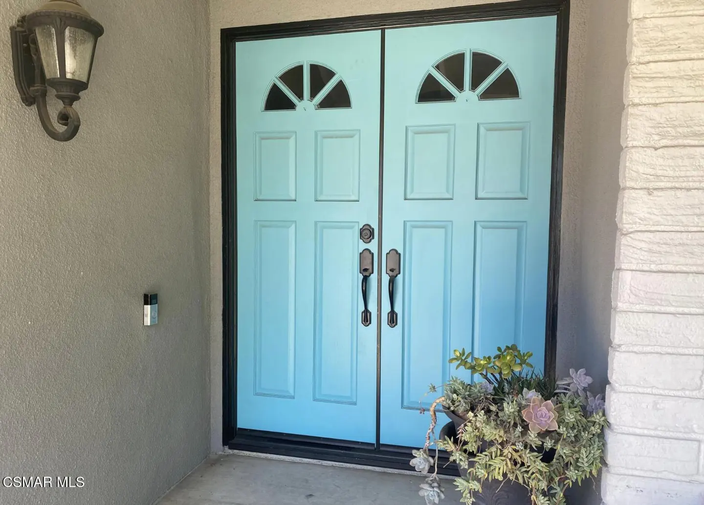 Exterior shot of a light blue double front door with black trim, a potted plant, and a wall-mounted light fixture.