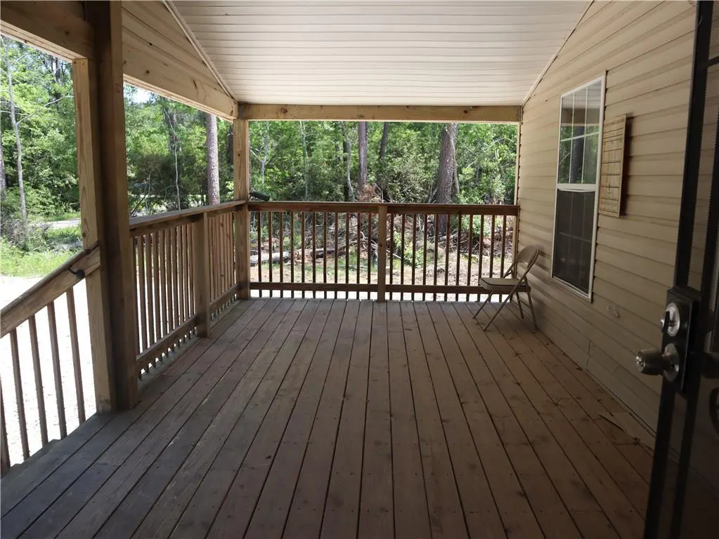 A wide, covered porch with wood railings overlooks a wooded area. A chair sits on the wood plank floor near a window.