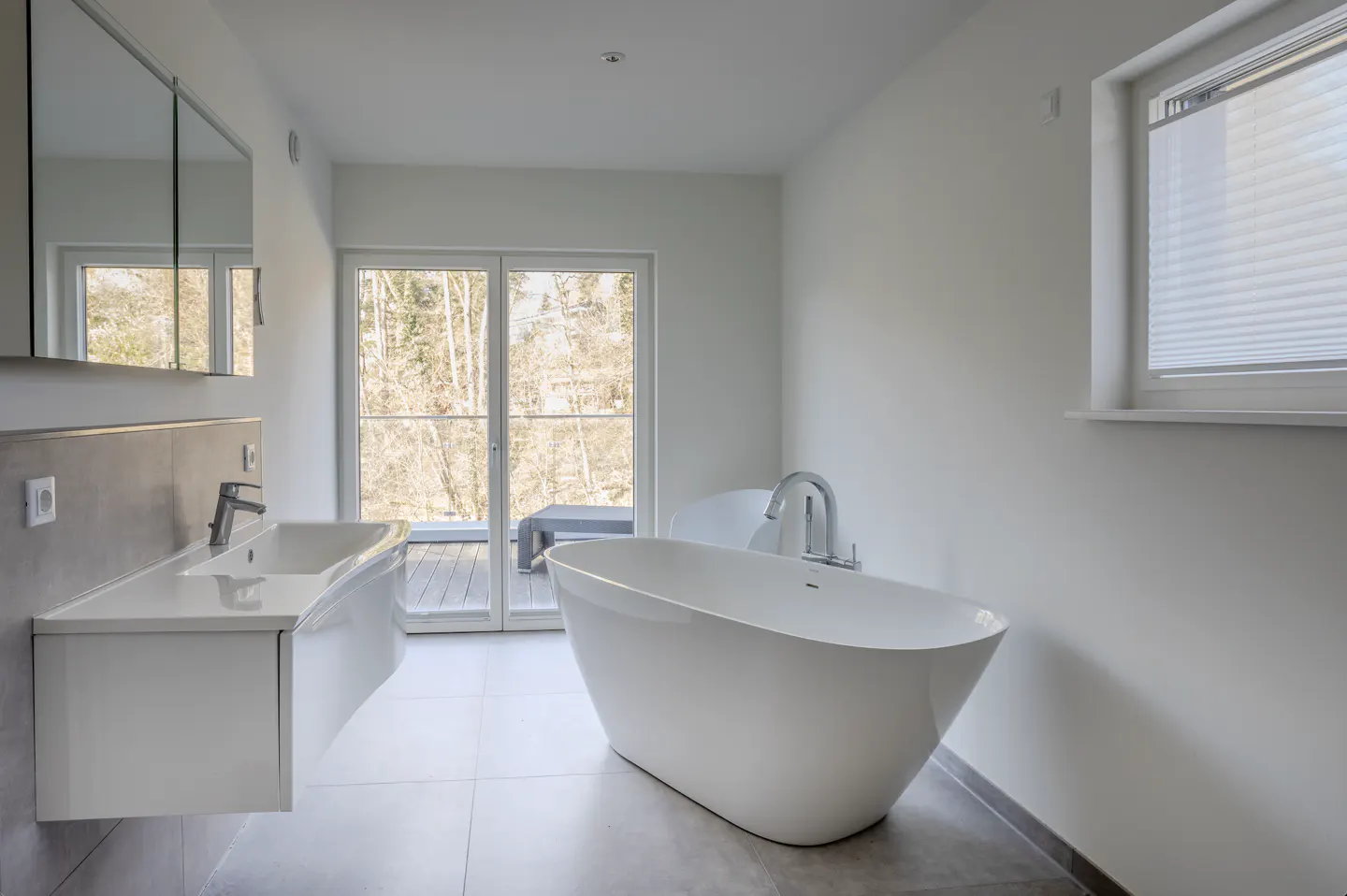 Bright bathroom with a white freestanding tub, modern sink, gray tile floor, and sliding glass doors to a balcony.