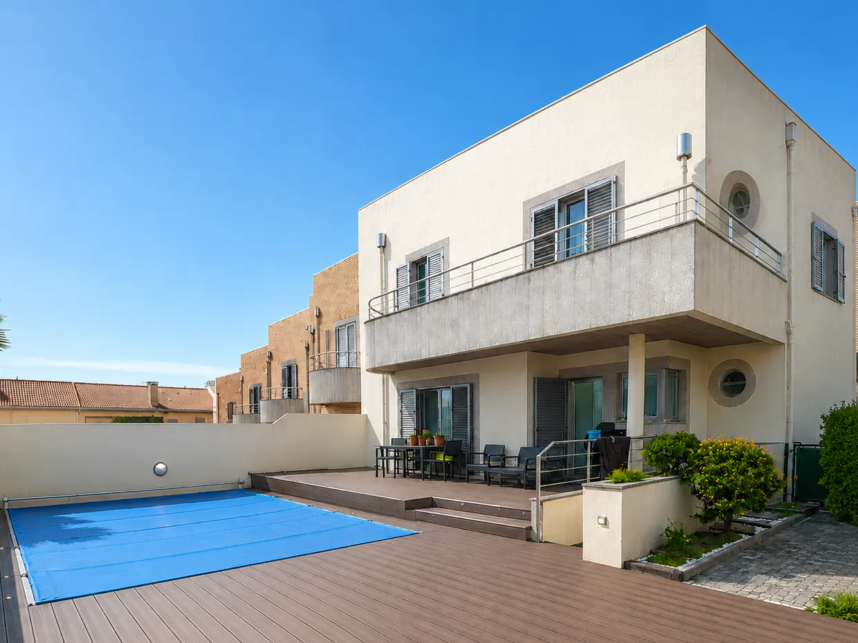 Exterior view of a two-story cream house with a balcony, a brown deck, and a blue covered pool under a clear blue sky.