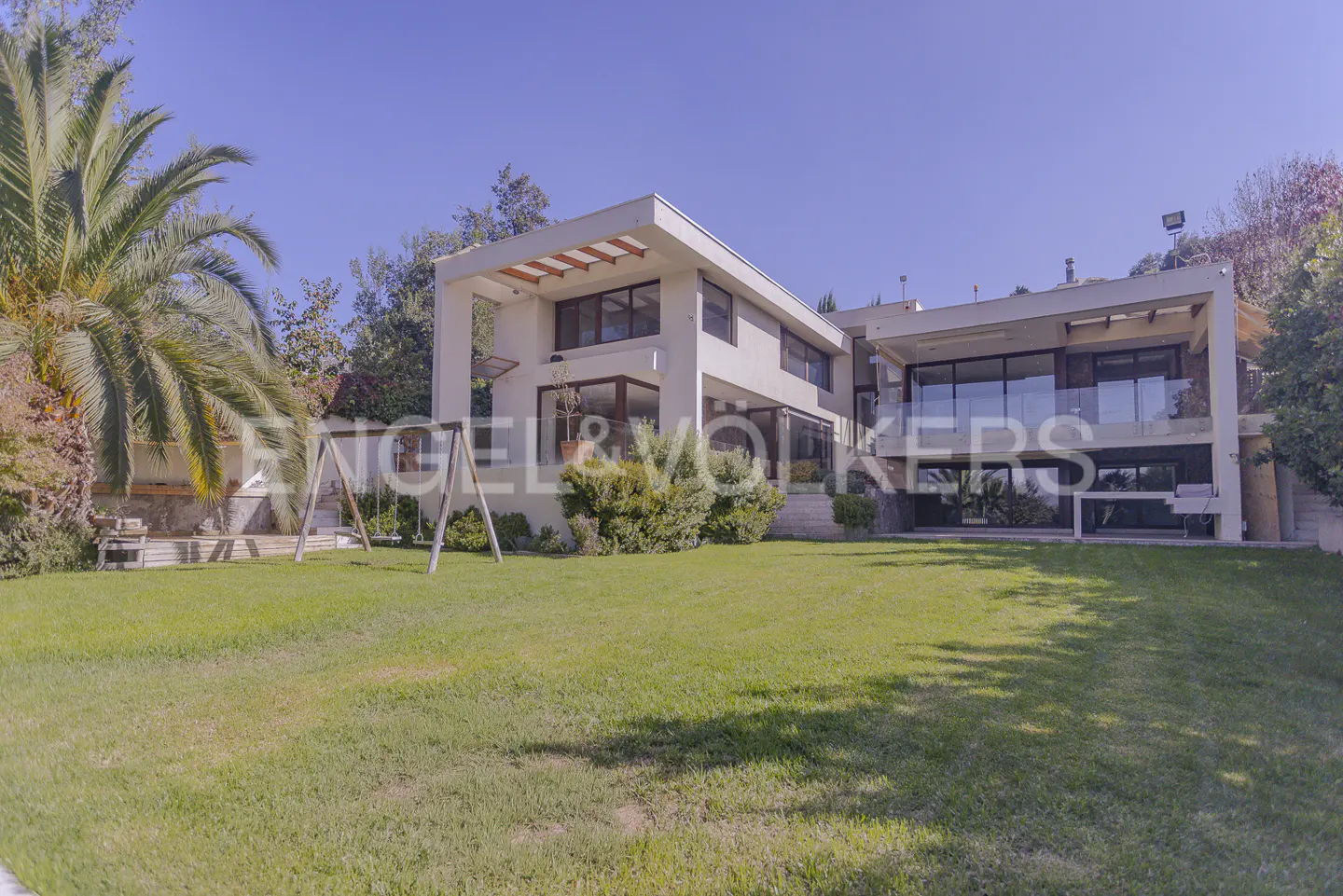 Modern two-story house with large windows and a green lawn. A swing set and palm tree are in the yard.