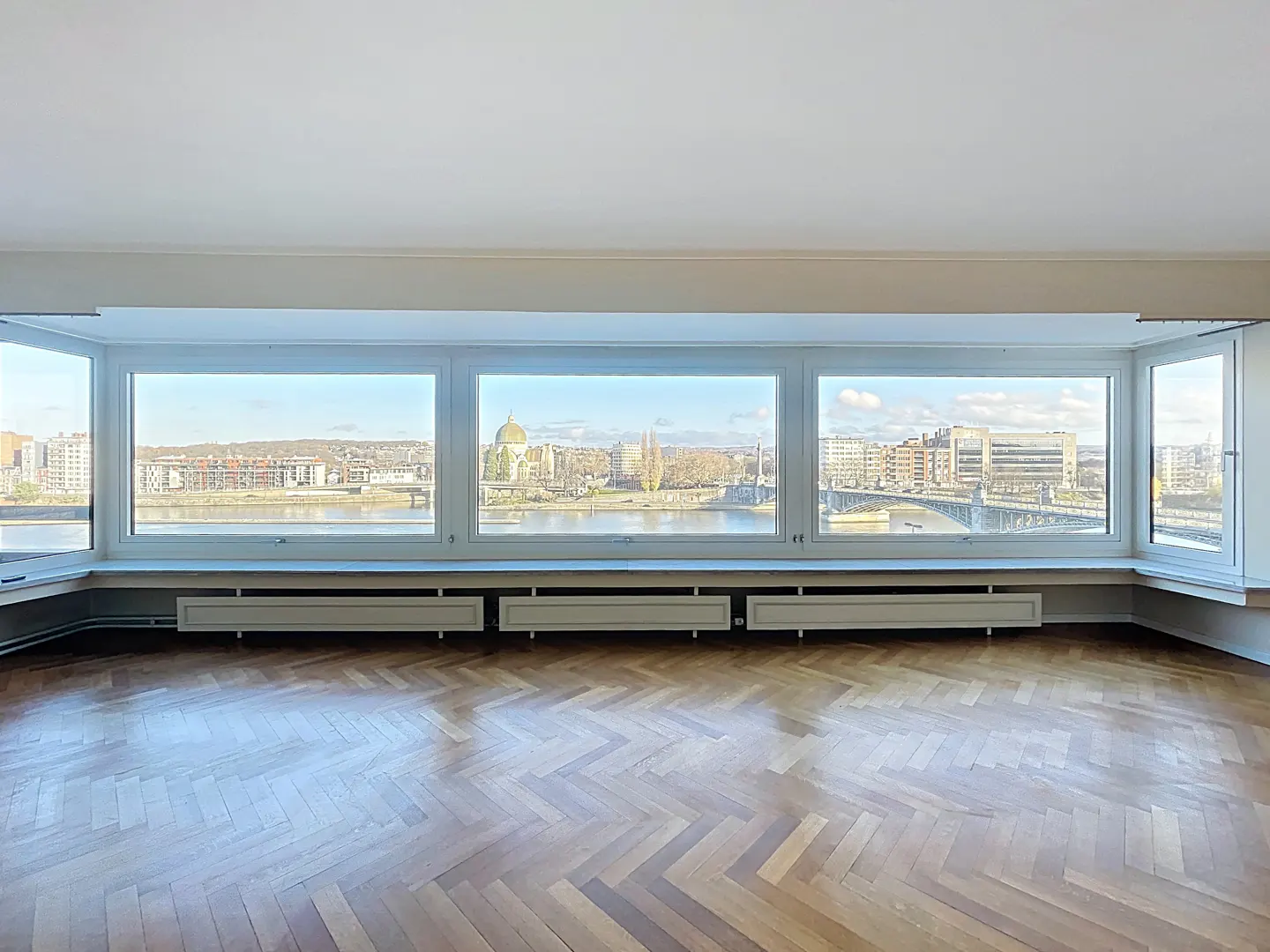 Bright, empty room with herringbone wood floor and large windows. Cityscape view includes a river, bridge, and buildings.