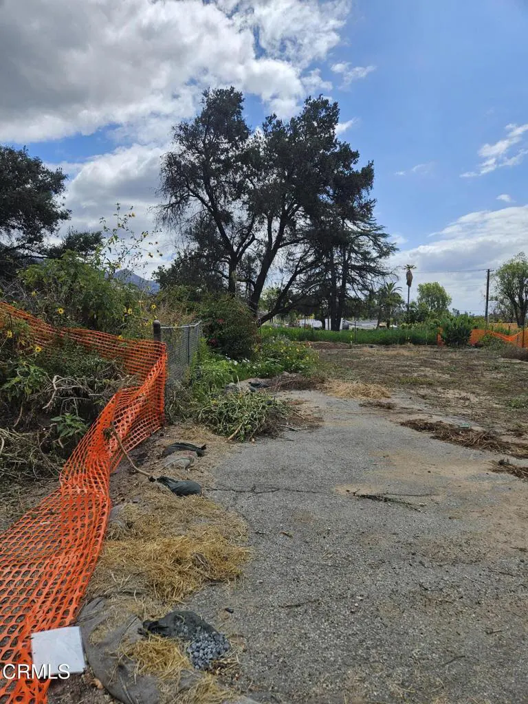 Vacant lot with cracked asphalt, orange safety fence, and trees under a cloudy blue sky.