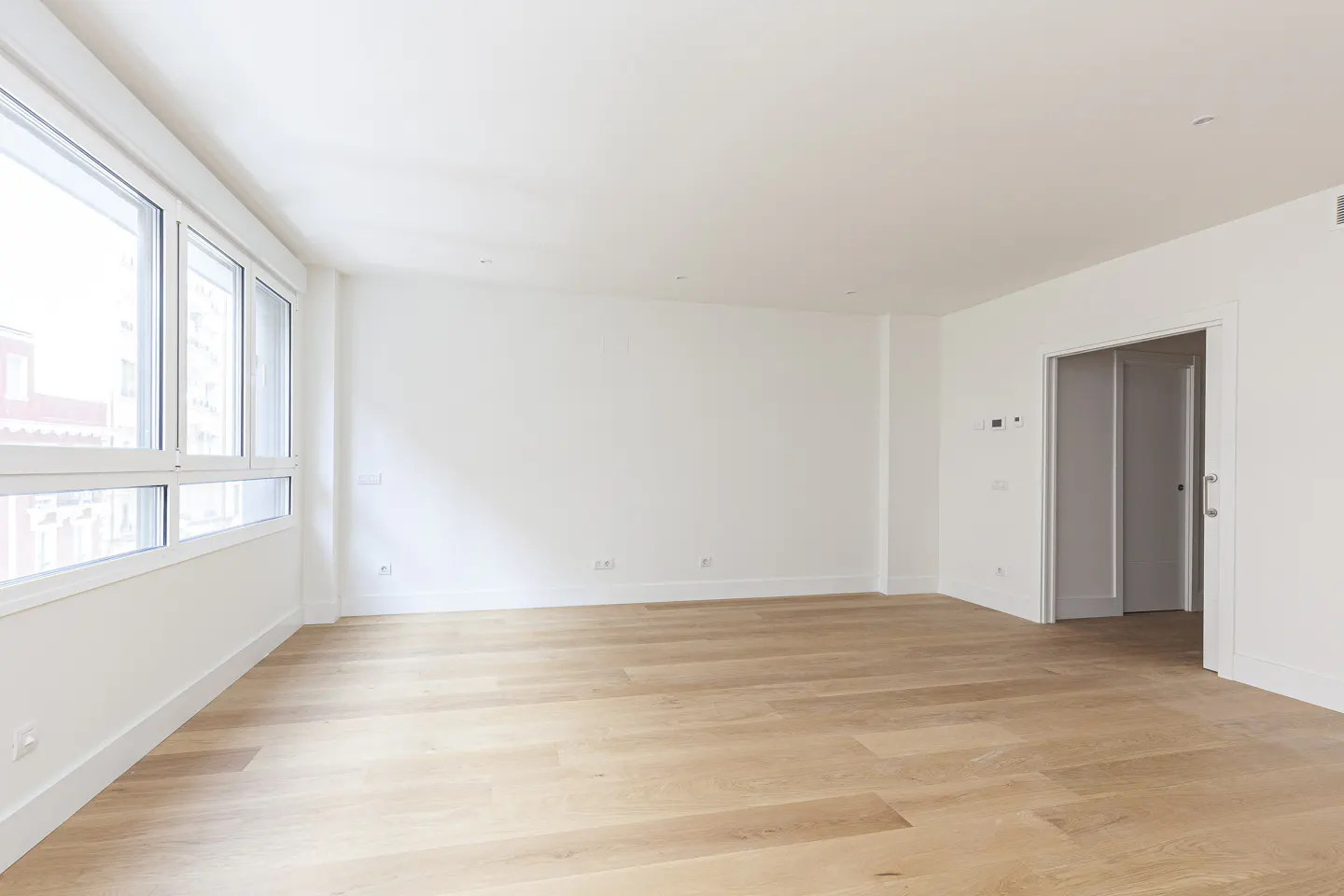 Bright, empty room with light wood floors, white walls, and large windows. A doorway is visible on the right.