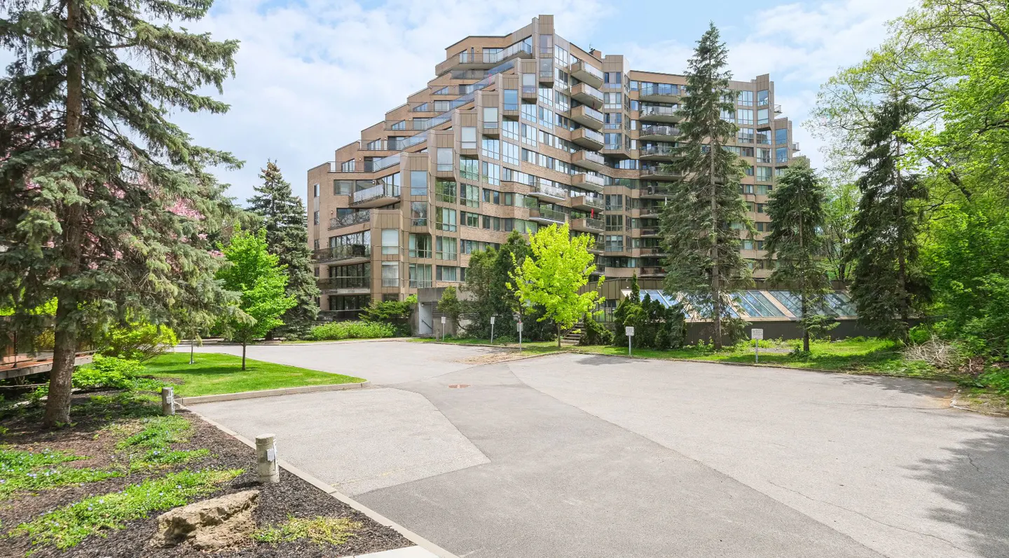 Exterior view of a modern beige condo building with balconies, surrounded by green trees and a paved parking area.