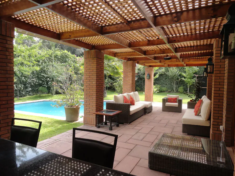 Outdoor patio with brick columns, a wooden lattice roof, and wicker furniture. A pool and green lawn are visible in the background.
