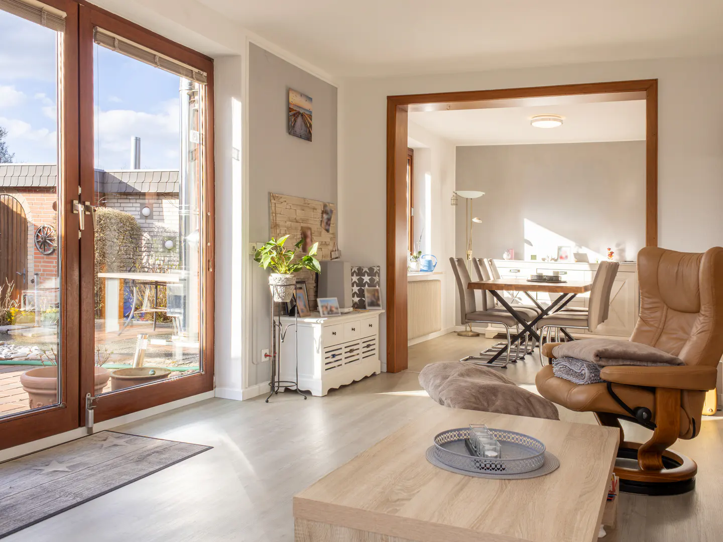Bright living room with wood-framed glass doors to a patio, a tan leather recliner, and a dining area visible through a wood-framed opening.