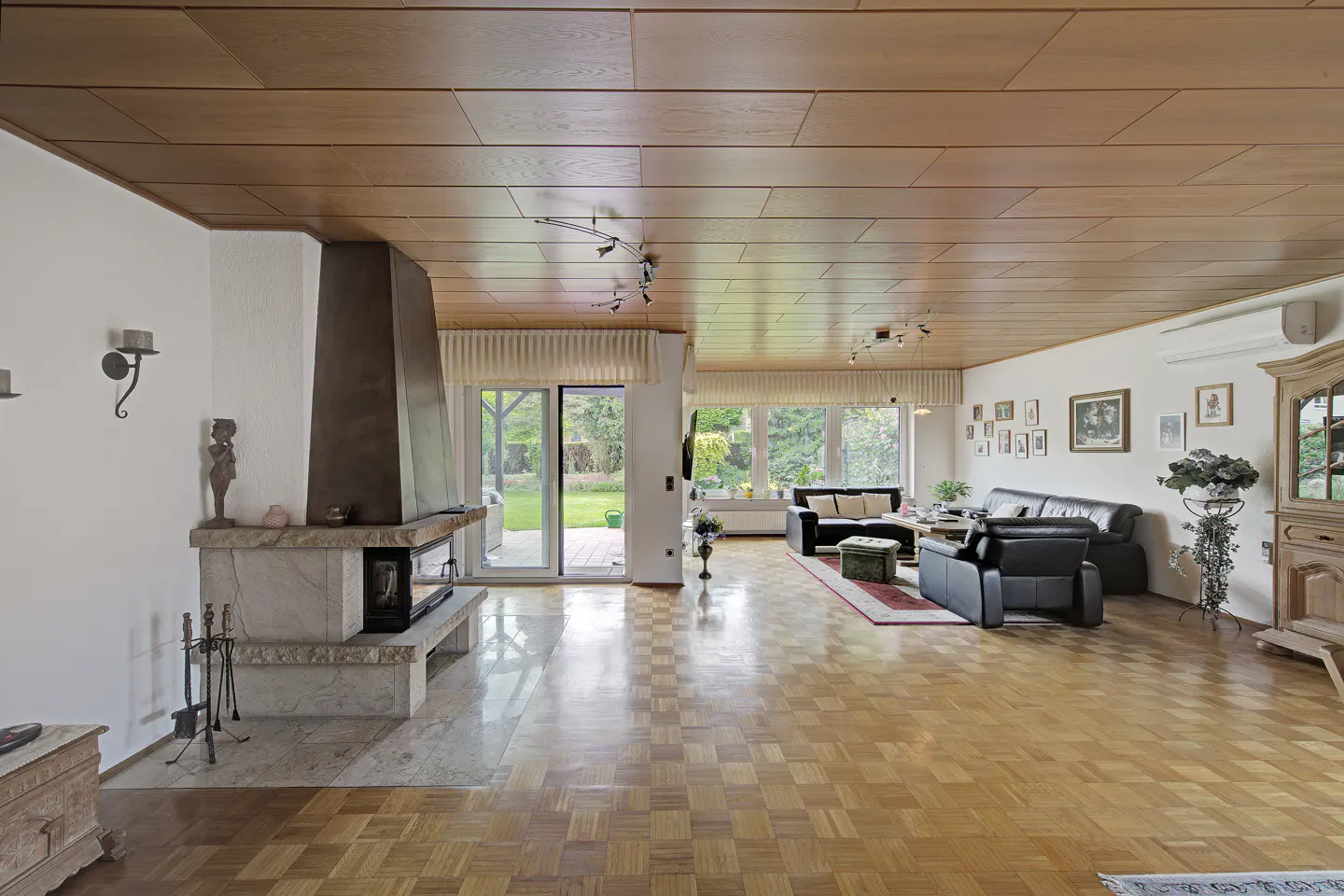 Spacious living room with wood-paneled ceiling, parquet floors, stone fireplace, and black leather furniture.