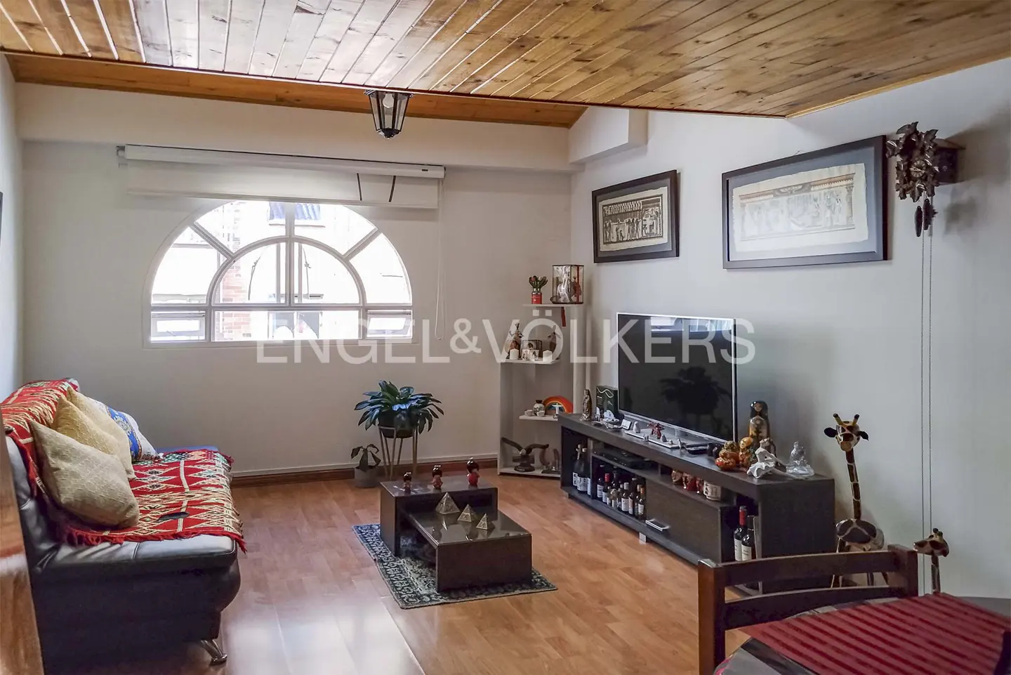 Living room with wood floors, a black sofa with a red blanket, and a TV on a dark wood stand. A round window lets in natural light.