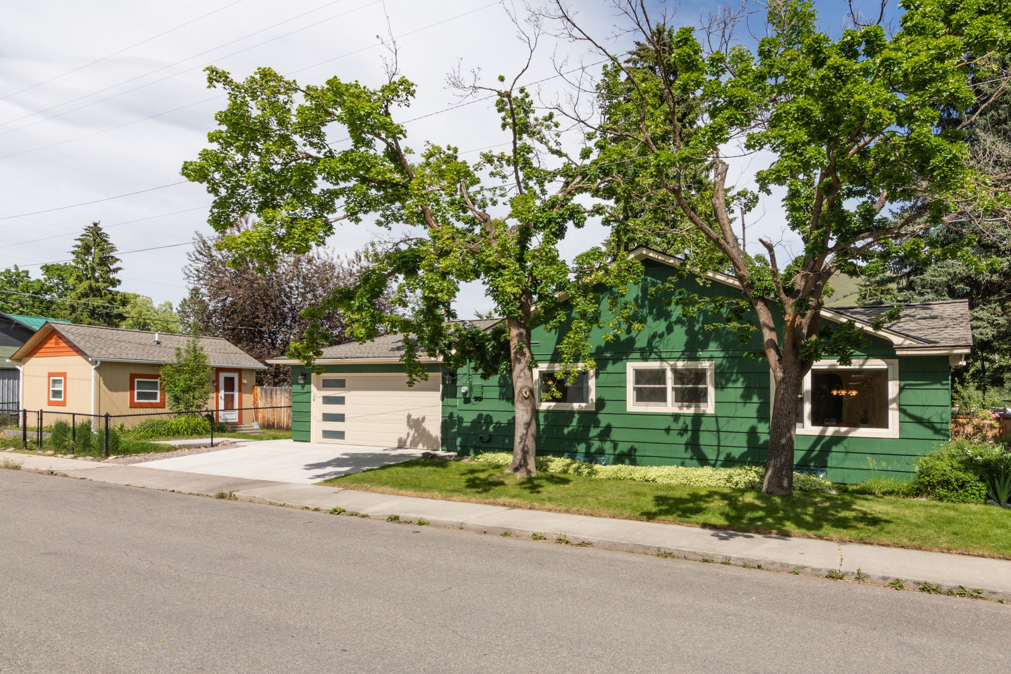 A green house with a tan garage and a smaller tan house in the background, viewed from a residential street.