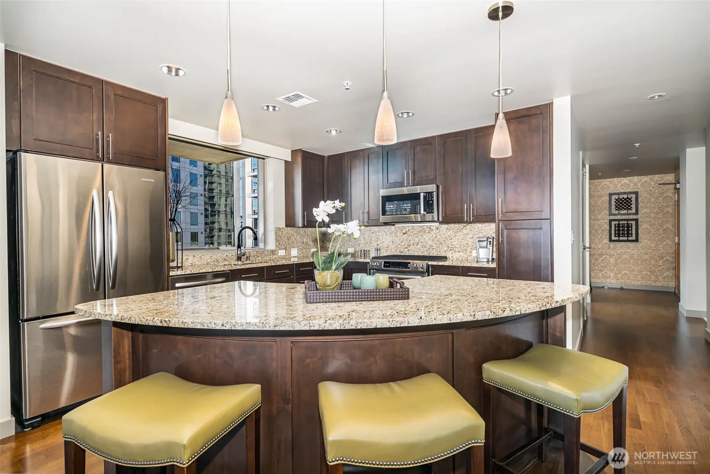 A modern kitchen with dark wood cabinets, granite countertops, and stainless steel appliances. Three pendant lights hang over a curved island with olive green stools.