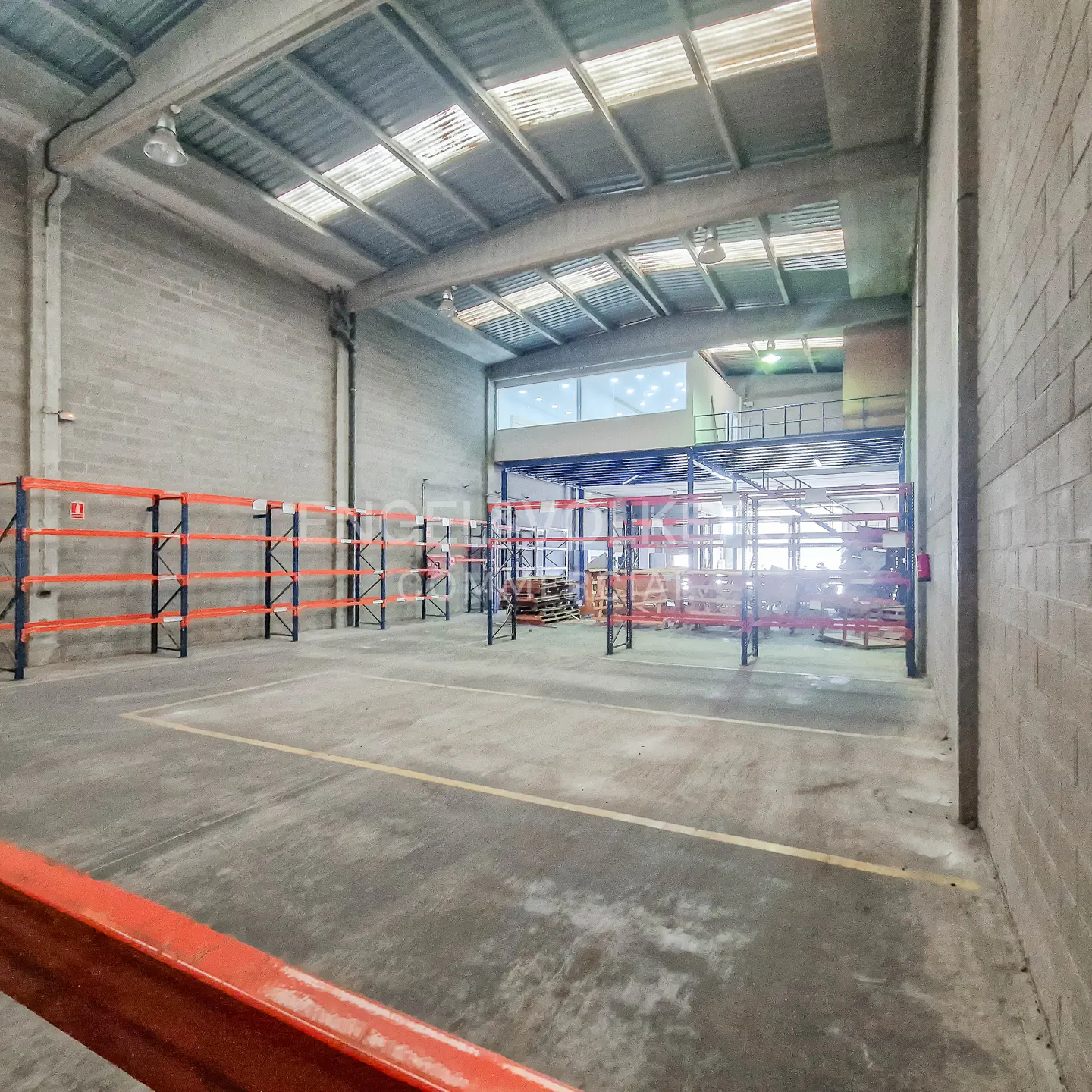 A large, empty warehouse with orange and blue metal shelving units. The floor is concrete with yellow lines. A metal roof and skylights provide natural light.