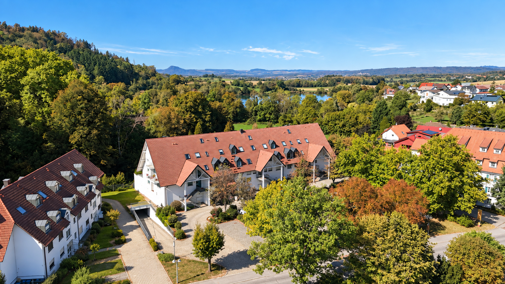 Aerial view of white apartment buildings with red roofs, surrounded by lush green trees and a distant lake under a blue sky. [AI-generated text]
