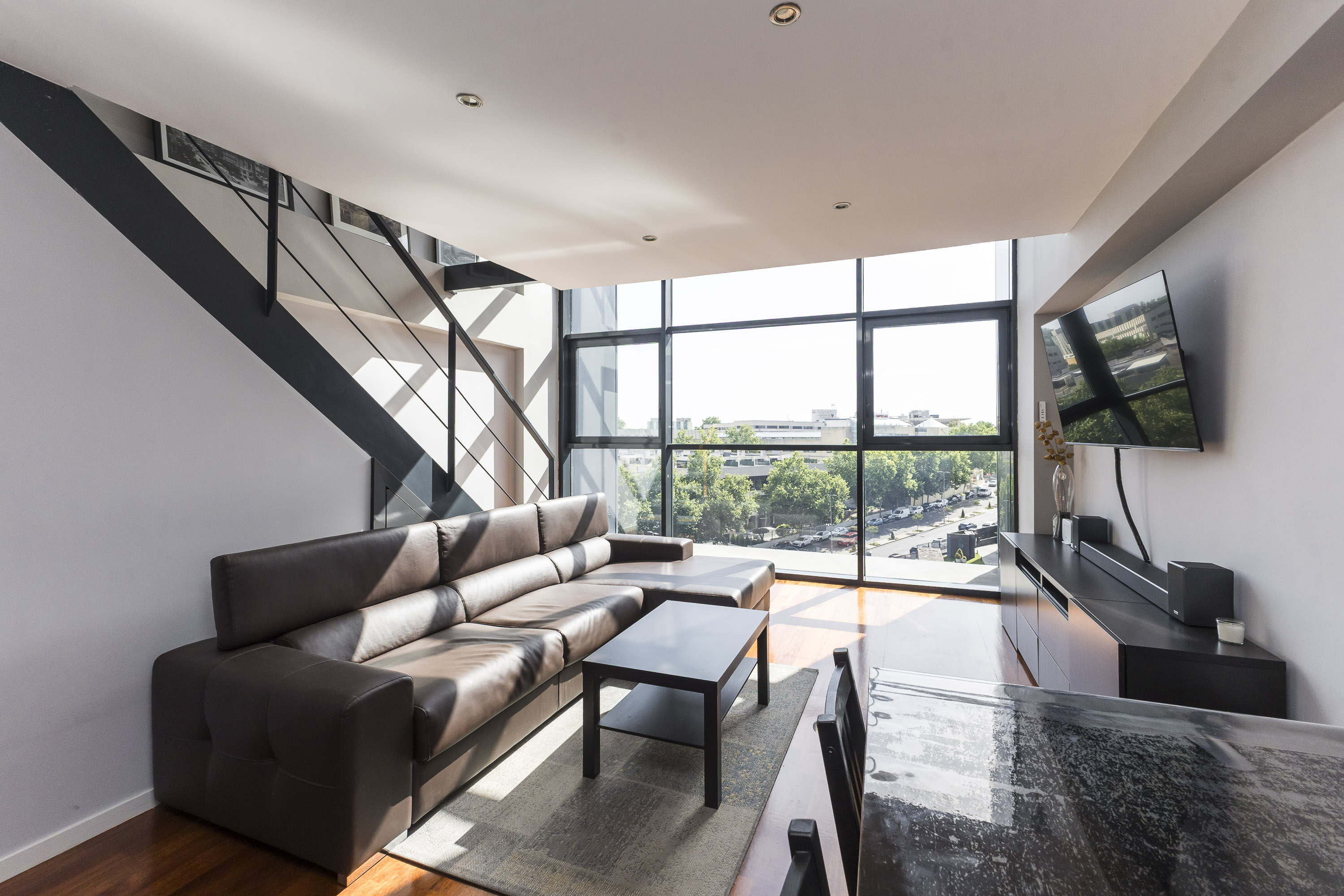 Modern living room with a brown leather sectional, coffee table, wall-mounted TV, and large windows overlooking a city.