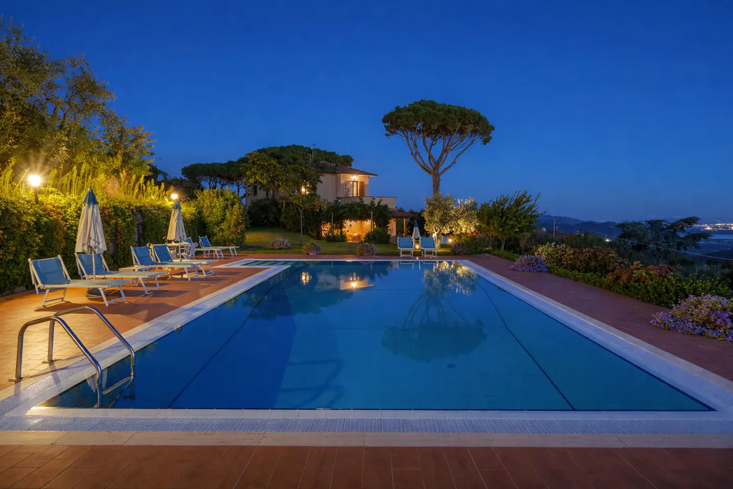 Outdoor pool at dusk with lounge chairs, umbrellas, and a house in the background. The pool reflects the lights.