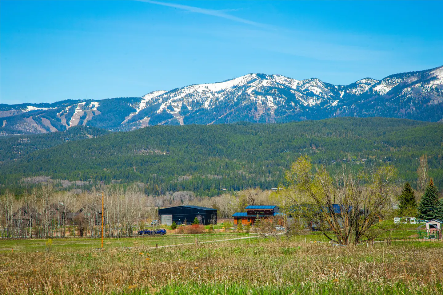 Landscape view of buildings in a field with snow-capped mountains and a blue sky in the background.